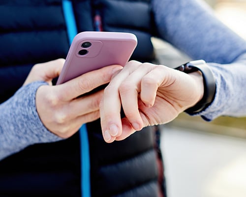 Woman in sports clothing looking at smartwatch and holding smart phone in her other hand, outdoors.