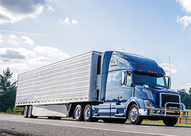 Blue semi truck hauling silver refrigerator semi trailer on a road in the summer road with forest and fields on the sides and blue sky and clouds above.
