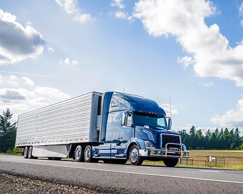 Blue semi truck hauling silver refrigerator semi trailer on a road in the summer road with forest and fields on the sides and blue sky and clouds above.