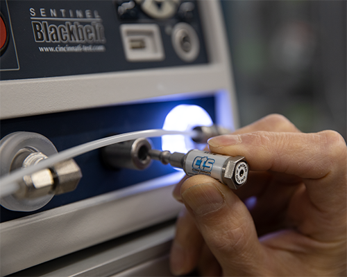 Hand shown inserting a CTS leak standard into the front of a leak test instrument in a factory.