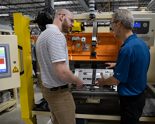 Two men standing in front of a leak test station on a factory floor. Man on left is looking at person on right who is explaining how it works.