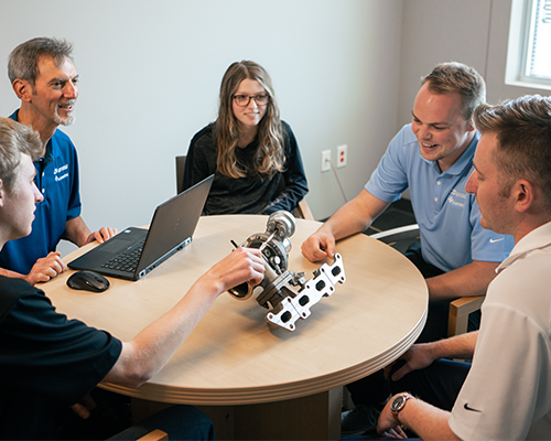 Four men and a woman sitting at a circular table in a small boardroom discussing how to leak test a metal part. One man is using a laptop, two are touching the metal assembly that is on the table. 