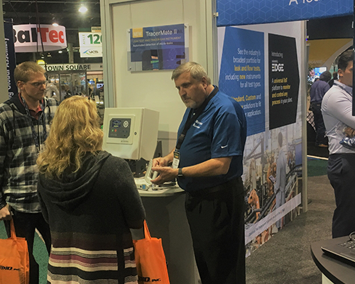 CTS employee in blue shirt standing in front of TracerMate II instrument on a pedestal speaking to a male and female attendee.