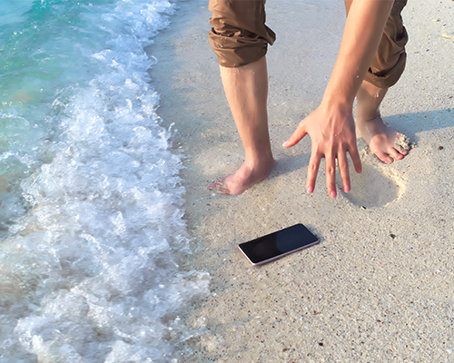 View of two feet standing on sand with hand reaching down to pick up cell phone which is near the edge of the water.