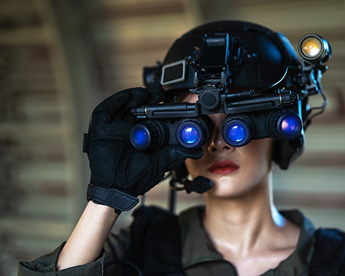 Female soldier wearing helmet using four-eyed night vision goggles in dark background.