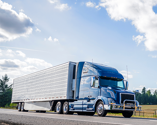 Blue semi truck hauling refrigerator semi trailer on a road in the summer road with forest and fields on the sides and blue sky and clouds above.